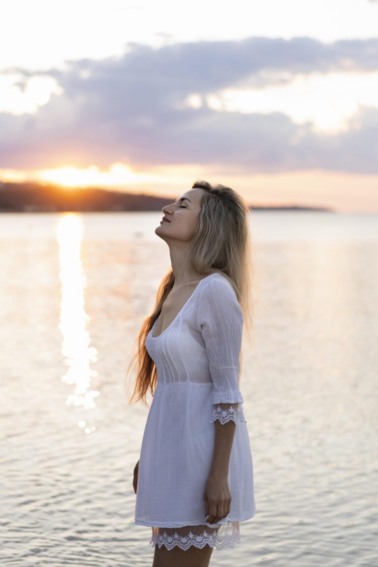 Jeune femme regardant le ciel au coucher du soleil au bord de l’eau, évoquant la quête de sens et le chemin de vie.