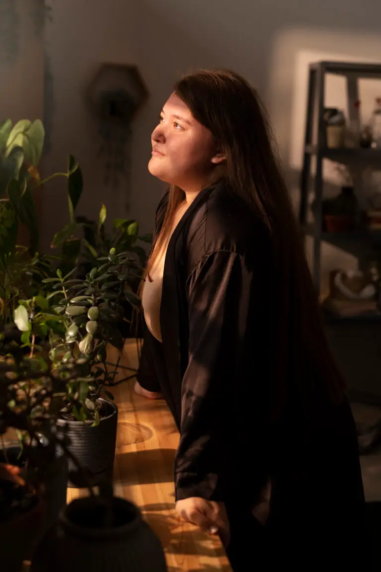 Jeune femme de profil regardant au loin, mains posées sur une table avec des fleurs en pot, baignée par la lumière du soleil, symbolisant le rapport à soi et aux troubles du comportement alimentaire