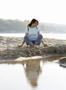 Jeune femme assise au bord d’un plan d’eau, observant son reflet brouillé dans l’eau