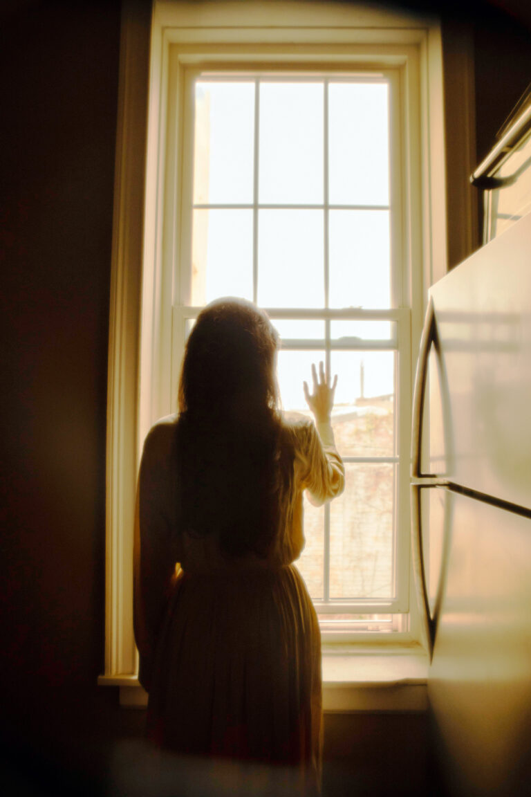 Jeune femme de dos, la main posée sur une fenêtre ouverte sur l’extérieur, dans une cuisine plongée dans la pénombre.